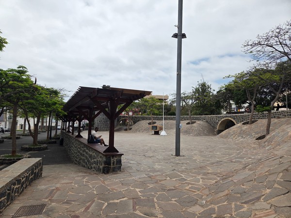 Plaza urbana en Adeje con pavimento de piedra, pérgola de madera con bancos y pequeños árboles, junto a un arco de mampostería en un día nublado.