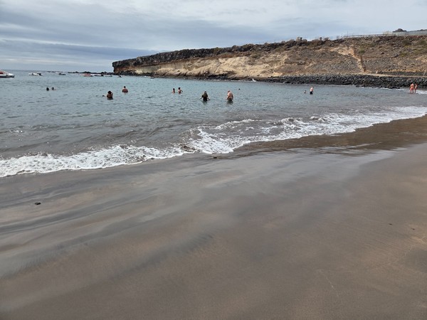 Orilla de Playa El Puertito con arena oscura, varias personas bañándose y acantilados claros al fondo en un día nublado.