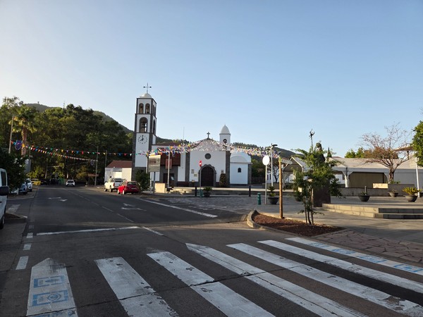 Plaza central de Santiago del Teide con iglesia blanca, torre con reloj y banderines de fiesta, rodeada de montes verdes.